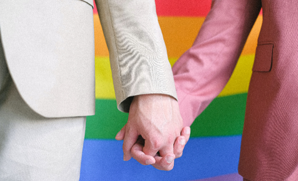 Dos personas tomadas de la mano frente a bandera LGBT